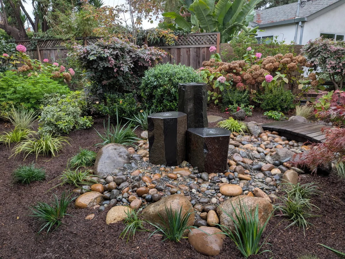 Three basalt column fountains flowing into river cobble, surrounded by lush garden planting